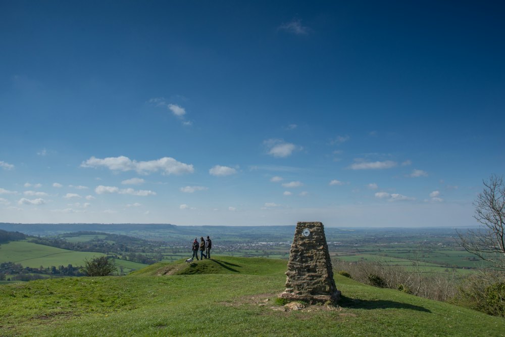 Walkers beside a stone trig point on a Cotswold hilltop, with the vale beyond