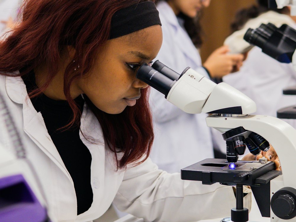 A student in a lab coat looking closely through a microscope